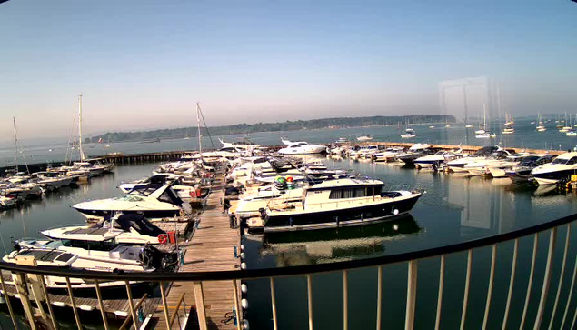 A view of a marina with numerous sailboats and motorboats docked along wooden piers. The water is calm and reflects the boats, and the sky is clear with no clouds. In the background, a distant shoreline with some low hills is visible. The photo is taken from behind a curved railing in the foreground.