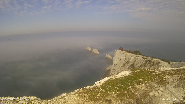 A panoramic view of dramatic white chalk cliffs rising sharply above the sea, with their edges highlighted by sunlight. Mist or fog hovers over the ocean below, partially obscuring the water and creating a misty atmosphere. A small building is situated on top of the highest point of the nearest cliff. The sky above is partly cloudy with touches of blue. The landscape appears rugged and remote, capturing a scenic coastal scene.