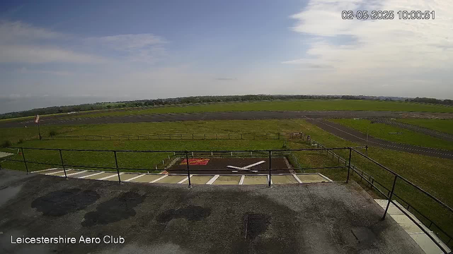 A wide view of an airfield with a helicopter landing pad in the foreground, surrounded by grass and fencing. The landscape extends into open fields and a distant horizon under a partly cloudy sky. The timestamp in the upper right corner reads "02-05-2025 10:00:51" and the bottom left corner states "Leicestershire Aero Club."
