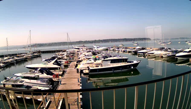 A marina with numerous boats and yachts docked along wooden piers extending into calm water. The sky is clear with a soft gradient from blue to a pale hue near the horizon. A metal railing curves along the foreground, partially framing the view. In the distance, there are more boats on the water and a low shoreline with trees.