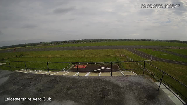 An outdoor view from a webcam shows a wide open grassy area with a runway or track in the distance under a cloudy sky. There is a metal railing in the foreground, and a small parking area or pit lane with white lines and a painted handicap symbol. The timestamp in the top right corner indicates the date and time as February 5, 2025, at 11:00:47. The bottom left corner displays the text "Leicestershire Aero Club."