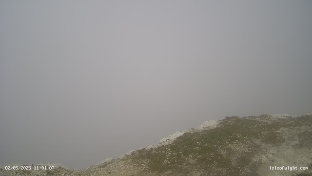 A foggy scene showing a rocky, grassy slope at the bottom with white mist obscuring the background. The date and time stamp in the lower left corner reads "02/05/2025 11:01:07," and a website URL "isleofwight.com" is in the lower right corner.