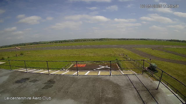 A wide view of an airfield under a partly cloudy sky. In the foreground, there is a parking area with white markings, a small fenced-off section with a shrouded object on the ground, and a parked vehicle. Beyond the parking area are grassy fields with a runway extending into the distance. To the left, there is a flagpole with a flag, and a low fence borders the airfield. The timestamp in the upper right corner reads "02-05-2025 11:59:54," and text at the bottom left says "Leicestershire Aero Club."