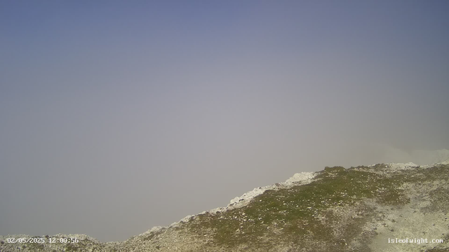 A viewpoint of a mountain landscape with rugged, rocky terrain in the foreground. The sky is clear and bright, with no visible clouds. The timestamp on the image indicates the date and time as February 5, 2025, at 12:00:56. The website "isleofwight.com" is displayed in the bottom right corner.