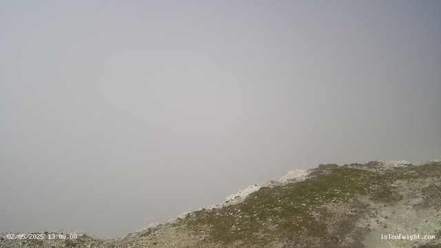 A rocky, grassy hillside with patches of white snow or chalk at its base. The sky is overcast and gray, with no visible clouds or sunlight. The image has a timestamp in the bottom left corner showing "02/05/2025 13:00:00" and a website watermark "isleofwight.com" in the bottom right corner.