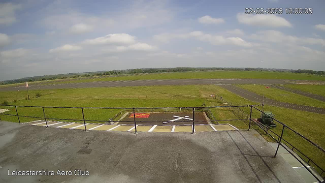 A view from a webcam at Leicestershire Aero Club showing a clear outdoor scene with a partly cloudy sky. In the foreground, there is an open space with a black railing and some parking spaces marked on concrete. To the right, a small vehicle or equipment is parked near a fenced area. Beyond the fencing, green grass fields stretch into the distance, along with a runway or taxiway area. The image is timestamped "02-05-2025 13:00:05" in the top right corner.