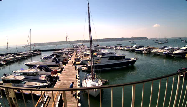 A marina filled with numerous boats and yachts docked along wooden piers, with the open sea in the background. The water is calm, and there is a clear sky overhead with a few scattered clouds. The photo appears to be taken from a balcony or deck with a railing in the foreground.