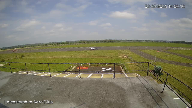 A view from a webcam at Leicesterhire Aero Club showing an airfield under a partly cloudy sky. In the foreground, there is a parking area with a marked helicopter landing pad and a small windsock. A small aircraft is visible on the runway in the distance, preparing for takeoff or landing. The surrounding landscape is mostly flat and grassy. The timestamp on the top right reads "02-05-2025 14:00:10".