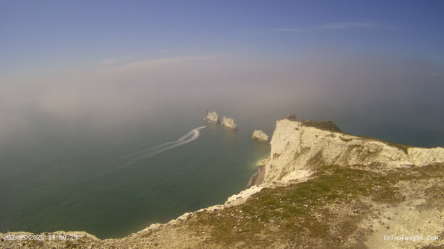 A view from a webcam shows white chalk cliffs extending into the ocean with some rocks and sea stacks in the water. The sky is partly cloudy with a gradient from blue to gray near the horizon. A boat is moving through the water, leaving a wake behind it. The timestamp in the lower left corner indicates the date and time as February 5, 2025, at 14:00.