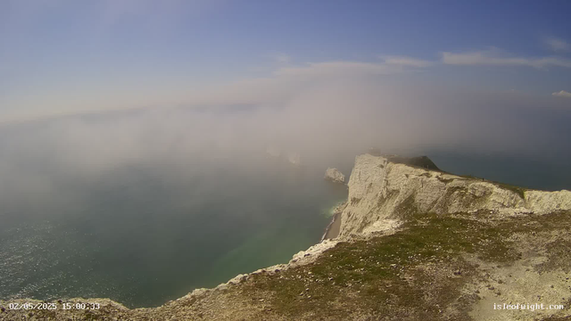 A view from a high cliff overlooking the ocean, with white chalk cliffs stretching along the coastline. There is some mist or fog partially obscuring the water and the distant horizon. The sky is mostly clear with a few clouds, and the cliffs are topped with green grass. The foreground shows a rocky, uneven surface at the edge of the cliff. The time stamp in the lower left corner indicates it was taken on February 5, 2025, at 3:00 PM.