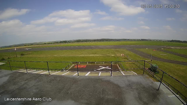 An outdoor view from a webcam at Leicestershire Aero Club shows a wide grassy airfield with a taxiway and runway extending into the distance under a partly cloudy sky. There is a small fenced area in the foreground with a few marked parking spaces and a run-in area, and a small vehicle parked on the right side. The timestamp in the top right corner indicates the date and time as February 5, 2025, 15:00:30.
