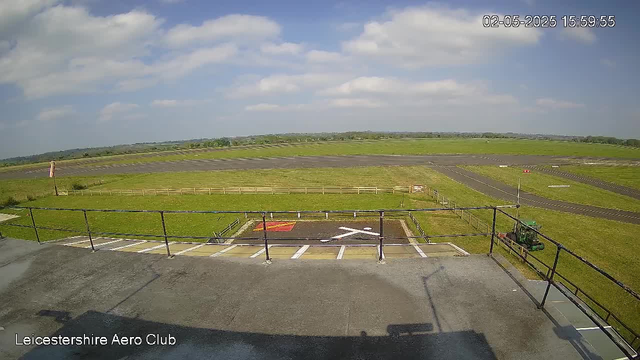 A view from a webcam at Leicestershire Aero Club showing an open grassy airfield with runways stretching into the distance under a partly cloudy sky. There is a small fenced area in the foreground with marked parking spaces, including a designated helicopter landing pad with an 'X' marking. A small vehicle is parked near the fence on the right side. The image includes a timestamp in the top right corner and the text "Leicestershire Aero Club" in the bottom left corner.