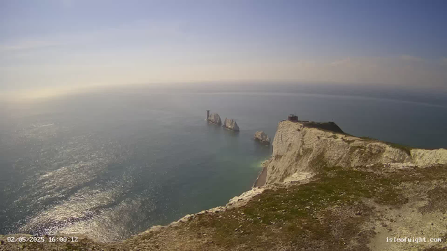 An aerial view of white chalk cliffs with patches of green grass on top, overlooking the ocean. In the distance, several large sea stacks rise from the water, with the tallest featuring a lighthouse. The sky above is mostly clear with a few wispy clouds, and the sunlight reflects off the water's surface. A small building is visible on the edge of the highest point of the cliffs. The timestamp in the bottom left corner indicates the date and time as February 5, 2025, at 4:00 PM.