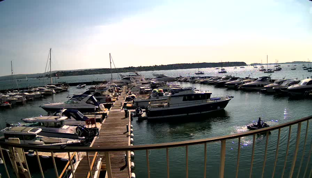 A marina filled with numerous boats and yachts docked along wooden piers. The water is calm, and the horizon features a distant coastline with a few structures. The sky is clear with a gentle light suggesting either early morning or late afternoon. A small boat with two people is moving across the water in the foreground.