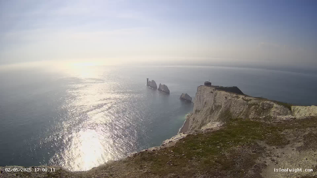 A view of rugged white cliffs extending into the ocean, with several large rock formations in the water near the base of the cliffs. The sky is clear with a soft, hazy sunlight reflecting off the calm sea. A small building or structure is perched on the edge of the cliff. The photo appears to be taken in the late afternoon or early evening.