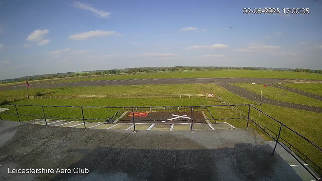 An outdoor view from a webcam at Leicestershire Aero Club showing a clear sky with a few scattered clouds. In the foreground, there is a small parking area with white lines, a red and yellow marked spot, and a black railing. Beyond the parking lot, there is a green grassy field with a narrow paved runway or track running through it. The horizon shows a distant line of trees and land, under a bright daylight sky. The timestamp on the image reads "02-05-2025 17:00:35".