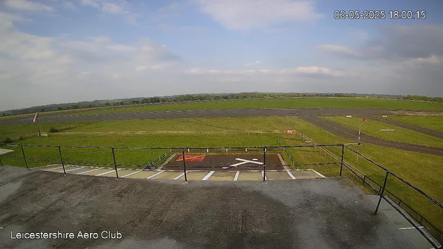 A view from a webcam at Leicestershire Aero Club showing a grassy airfield with runway and taxiway extend into the distance under a partly cloudy sky. In the foreground, there are parking spaces, a railing, and a small flight safety sign. The timestamp in the top right corner indicates the date and time as May 2, 2025, 6:00 PM.