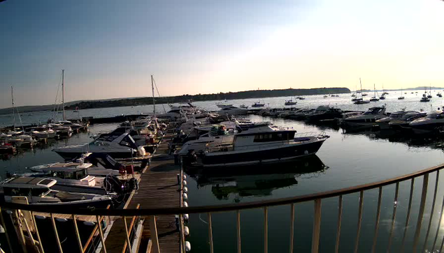 A marina filled with numerous boats and yachts docked along wooden piers, with calm water reflecting the vessels. In the background, there are more boats floating on the water, with a low shoreline and a clear sky overhead, suggesting late afternoon or early evening lighting.