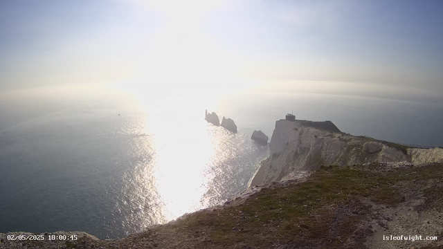 A view of white cliffs overlooking the ocean during sunset, with a lighthouse on top of one of the cliffs and several rocks protruding from the water. The sunlight is reflecting brightly off the sea, creating a glare that partly obscures the horizon. The date and time displayed are "02/05/2025 18:00:45," and there is a website URL "isleofwight.com" in the bottom right corner.