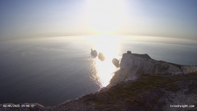 A view of white chalk cliffs overlooking the ocean at sunset. The sky is clear with a bright sun low on the horizon, reflecting off the water's surface. Three sea stacks are visible near the base of the cliffs, with a small structure and a lighthouse atop the cliffs. The image includes a timestamp with the date "02/05/2025" and the time "19:00:47," along with the website "isleofwight.com" in the bottom right corner.