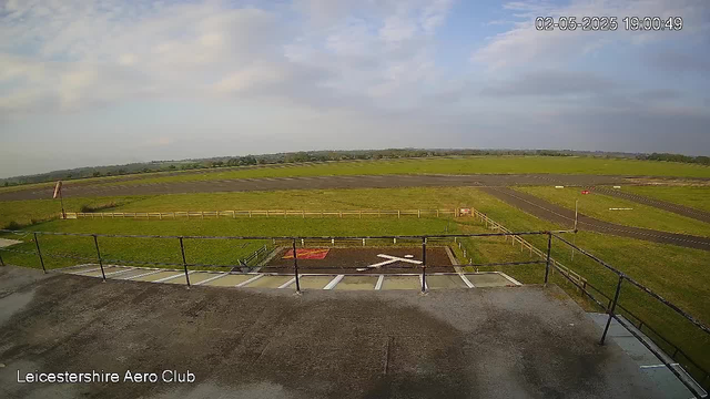 An outdoor view from a webcam at Leicesterhire Aero Club shows a clear sky with some scattered clouds, a grassy area enclosed by a low fence, and a paved runway extending into the distance. There is a section of parking or loading area in the foreground with marked lines, and a windsock on the left side of the image. The timestamp in the top right corner shows the date as May 2, 2025, and the time as 19:00:49.