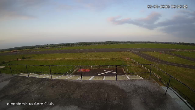 An outdoor view from a webcam at Leicestershire Aero Club, showing a cloudy sky with a few pink-tinged clouds at sunset or sunrise. In the foreground, there is a flat concrete rooftop with a black railing and a small marked area. Beyond the rooftop, there is a grassy field with a fence and a narrow paved runway stretching into the distance. The landscape is mostly flat with some gentle rolling hills on the horizon. The timestamp in the upper right corner shows the date and time as February 5, 2025, at 8:00 PM.