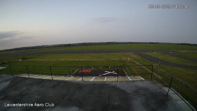 An outdoor view of a small aerodrome with a helicopter landing pad in the foreground. The landing pad has a white cross and a red marking, surrounded by a black border. There is a black railing around the edge of a concrete platform above the landing area. Beyond the landing pad, there is a flat grassy area, a narrow runway, and an open landscape with a distant tree line under a partly cloudy sky. The timestamp in the top right corner reads "02-05-2025 21:00:40." The bottom left corner has the text "Leicestershire Aero Club."