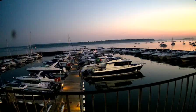A marina at dusk with numerous sailboats and motorboats docked along wooden piers. The water is calm, reflecting the boats and the gradually darkening sky, which has a soft gradient of colors from light pink to blue. A curved railing is visible in the foreground.