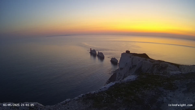 A view of white chalk cliffs at sunset, with a small building and a lighthouse on top of the cliffs. The sea below is calm, reflecting the colors of the sky, which transitions from pale blue to warm orange and yellow near the horizon. A narrow pathway runs along the edge of the cliffs. The timestamp in the bottom left corner reads "02/05/2025 21:01:05," and the bottom right corner has the website "isleofwight.com."