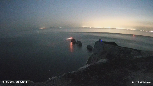 A view of a coastal scene at dusk or dawn with the sky transitioning from dark to light. The water is calm and stretches out towards the horizon, where distant city lights create a faint glow. Several rocks are visible emerging from the water near the foreground, and a small light is situated on one of the rocks. The photo has a timestamp in the lower left corner showing "02/05/2025 21:59:43" and a website address "isleofwight.com" in the lower right corner.