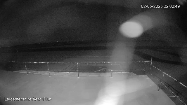 A black-and-white image of an outdoor sports field at night, with a metal fence surrounding it. The sky is dark, and a bright, large, round object, possibly the moon, is visible in the upper right part of the image, casting a faint glow. The top right corner displays the date and time: "02-05-2025 22:00:49."