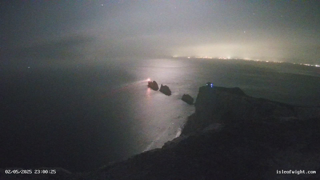 A nighttime view of a rocky coastline with large boulders protruding from the water. In the background, there are distant city lights along the horizon. The sky is mostly cloudy with a few visible stars. The timestamp on the image reads "02/05/2025 23:00:25," and the website "isleofwight.com" is displayed in the bottom right corner.