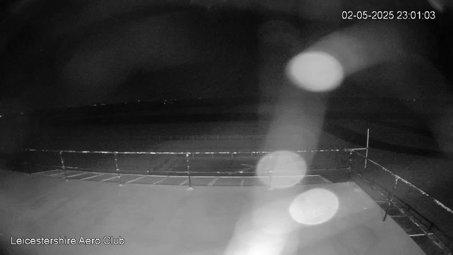 A black-and-white image taken by a webcam showing an empty outdoor area at night with a metal fence along the edge. The scene is dark, and the sky is mostly black with some faint distant lights visible on the horizon. The top right corner displays a timestamp reading "02-05-2025 23:01:03." The bottom left corner contains the text "Leicestershire Aero Club." There are several bright, circular spots of light or glare across the image, likely caused by condensation or dirt on the camera lens.