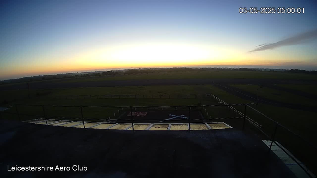 A wide outdoor view of a flat, open landscape at sunrise or sunset, with the sky showing a gradient of colors from yellow near the horizon to light blue overhead. The foreground includes part of a rooftop or platform with a railing, and the horizon stretches across the image with no prominent structures. The scene is peaceful and illuminated gently by the low sunlight. The image has a timestamp in the upper right corner reading "03-05-2025 05:00:01" and the bottom left corner displays the text "Leicestershire Aero Club."