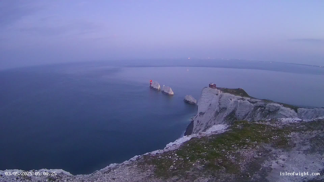 A view from a seaside cliff at dawn or dusk, showing white chalk cliffs and rock formations rising from calm blue-green waters. A lighthouse with a red light stands on a small island or rock in the water, and a buildings sit atop the cliffs. The sky is clear with soft pastel colors. The timestamp shows the date as May 3, 2025, at 5:00 AM. The website "isleofwight.com" is visible in the corner.