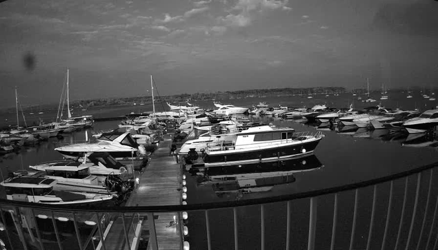 A black-and-white image of a marina with numerous boats docked along wooden piers. The boats vary in size, with some larger yachts and smaller vessels. The water is calm, reflecting the boats and the sky above, which has scattered clouds. A curved railing is visible in the foreground, indicating the photo was taken from a vantage point overlooking the marina.