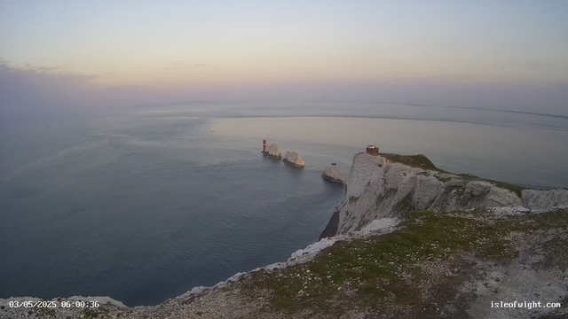 A view of white chalk cliffs overlooking the ocean at dawn. A lighthouse sits on top of the cliff, and several large chalk rock formations are visible in the water nearby. The sky is light with subtle colors, indicating early morning.