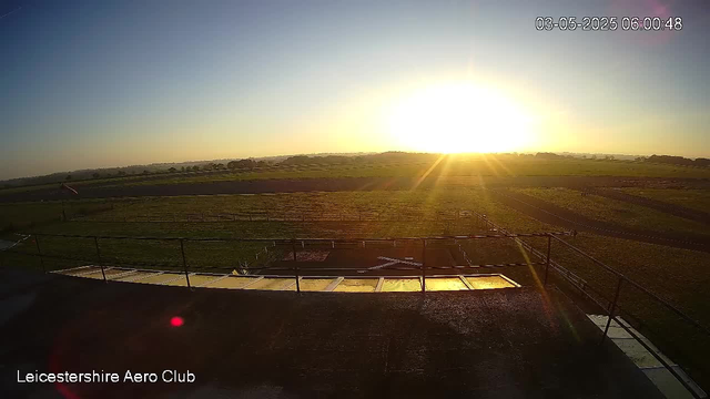A wide view of an open grassy field with a small dirt track and fencing in the foreground, illuminated by the bright, setting sun near the horizon. The sky is mostly clear with a subtle gradient from blue to warm tones near the horizon. The timestamp in the top right corner shows the date and time as May 3, 2025, at 6:00:48 AM. The bottom left corner contains the text "Leicestershire Aero Club."