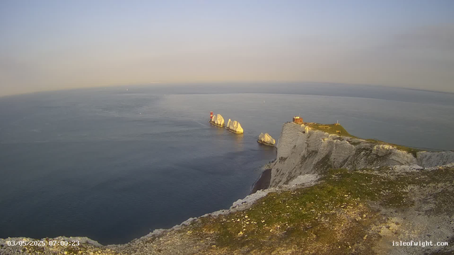 A panoramic view of white chalk cliffs with green grassy tops stretch along the coastline, overlooking the calm ocean waters. Large white rock formations emerge from the sea near the base of the cliffs. On the highest point of the cliffs, there is a small building and a lighthouse with red and white stripes. The sky is clear with a soft gradient from pale blue to light yellow near the horizon. The image has a timestamp in the bottom left corner and a website URL in the bottom right.