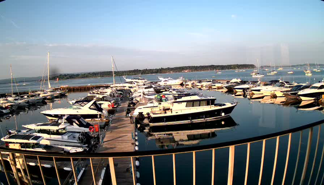 A marina filled with various boats docked along wooden piers, with calm water reflecting the boats. Several sailboats and motorboats are visible, with a distant shoreline and a cloudy sky in the background. A metal railing curves in the foreground, framing the scene.