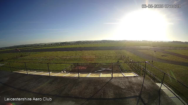 An outdoor view from a webcam at Leicesterhire Aero Club showing a grassy airfield with a few paved runways stretching into the distance under a bright, sunny sky. The sun is low on the horizon, casting strong sunlight and long shadows. In the foreground, there is a fenced area with some paved surfaces, and the background features open fields and a clear blue sky. The timestamp in the top right corner reads "03-05-2025 07:00:29."