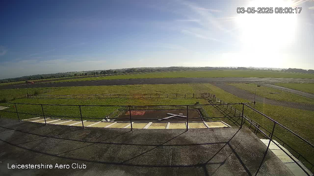A wide view of a grassy airfield under a clear blue sky with the sun shining brightly to the right. In the foreground, there's a fenced area with some white markings on the ground, possibly a small helicopter landing pad. Beyond that, the landscape extends to open fields and a runway. The timestamp in the upper right corner indicates the date and time as March 5, 2025, at 8:00 AM. The text "Leicestershire Aero Club" appears in the lower left corner.