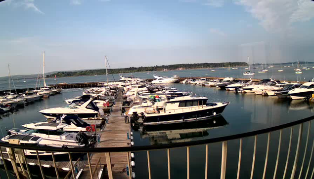 A marina filled with numerous boats docked along a wooden pier. The boats vary in size and shape, with some having cabins and others being open. Calm water reflects the boats, and in the background, there are more yachts anchored further out in the water. The sky is partly cloudy with patches of blue.