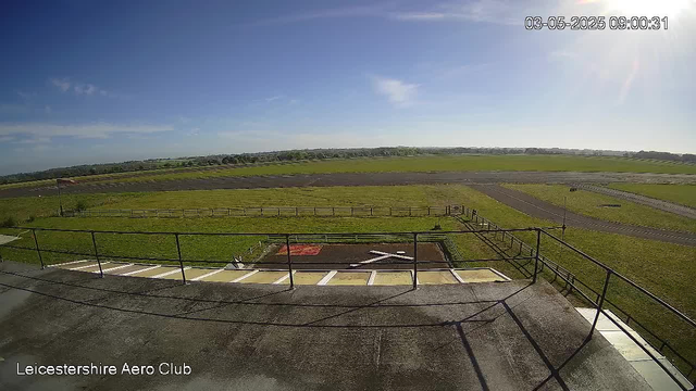 A view from a webcam showing the LeicesterShire Aero Club's outdoor area. In the foreground, there is a tarmac surface with a section of a helipad marked with a white "H". Surrounding the helipad is a metal railing. Beyond the railing, there are open grassy fields with runways and taxiways, and a clear blue sky with a few scattered clouds. The time and date stamp in the upper right corner indicates "03-05-2025 09:00:31".