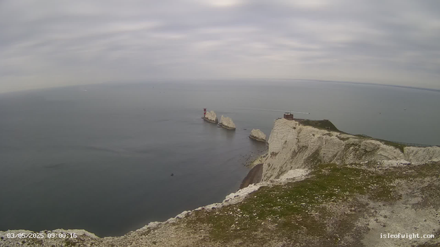 A view of white chalk cliffs extending into the sea, with three large sea stacks rising from the water in the distance. The sky is overcast with gray clouds, and the water appears calm. There is a small building or lighthouse on top of the cliffs near the right side of the image. The shoreline at the bottom is rocky with patches of grass. The image has a timestamp of "03/05/2025 09:00:16" in the lower left corner, and the website "isleofwight.com" is visible in the lower right corner.