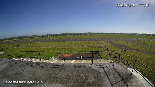 An outdoor view from a webcam showing a clear, sunny day at Leicestershire Aero Club. In the foreground, there is a paved area with parking spaces and a railing. Beyond that, there is a grassy field with some pathways and fences. The horizon displays a distant line of trees under a bright blue sky with no visible clouds. The date and time in the top right corner indicate it is 10:00 AM on May 3, 2025.
