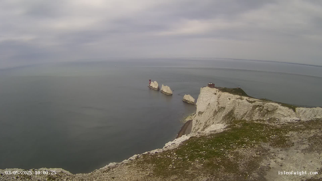 A panoramic view of white chalk cliffs rising steeply from the sea, with several large white rocks near the shoreline. There is a small red lighthouse on one of the rocks in the water. The sky is overcast with gray clouds, and the sea appears calm. A small building or observation point is situated on the top of the cliffs. The timestamp at the bottom left indicates the date and time as 03/05/2025 at 10:00:25.