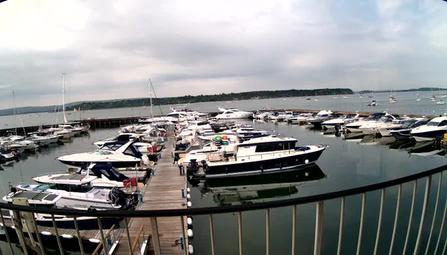 A view of a marina with numerous boats and yachts docked along a wooden pier. The water is calm, reflecting the boats and a cloudy sky overhead. The background features a distant shoreline with more boats floating in the open water.