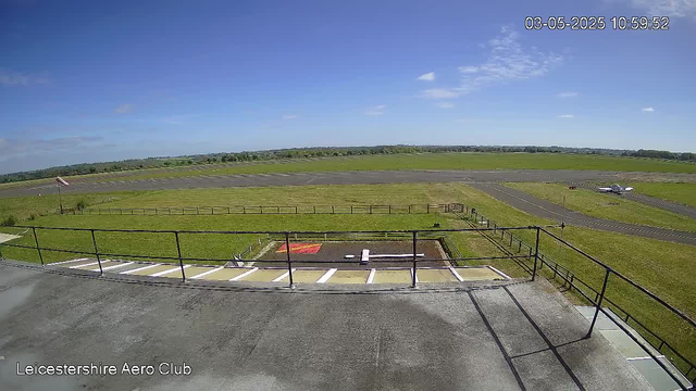A view from a webcam at Leicestershire Aero Club shows an airport runway with a small aircraft on the tarmac in the distance. The sky is clear with a few scattered clouds, and the surrounding area is green and open. There is a fence and staircase in the foreground. The timestamp in the top right corner indicates the date and time as March 5, 2025, at 10:59:52.