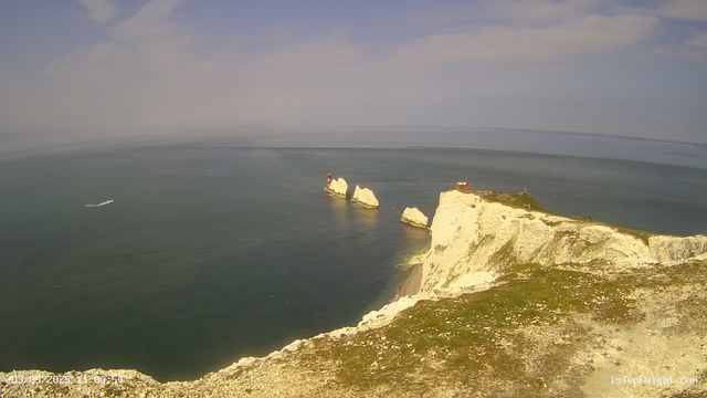 An aerial view showing white chalk cliffs along a coastline with a grassy top, a small building, and a lighthouse. The cliffs overlook a calm, greenish-blue sea with a boat sailing nearby. The sky is partly cloudy with patches of blue. The date and time are displayed at the bottom left corner.